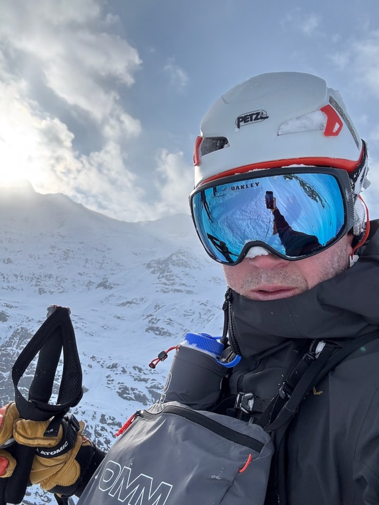A climber wearing a white helmet and mirrored goggles stands on a snowy mountain peak, holding climbing gear and a water bottle, with a cloudy sky in the background.