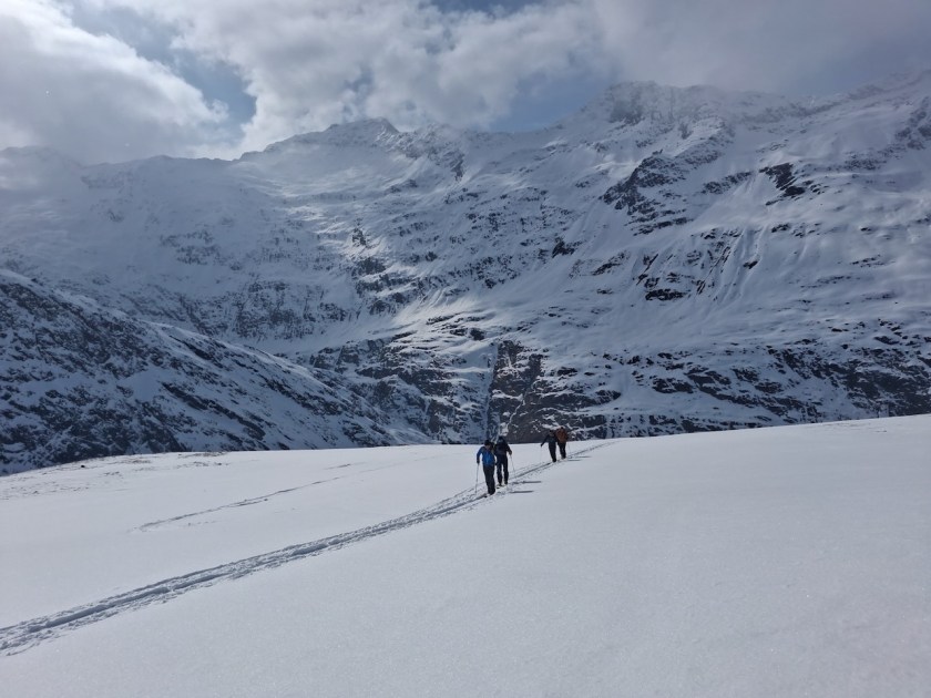 Group of hikers walking on a snow-covered mountain landscape with tall, snow-capped peaks and a cloudy sky.
